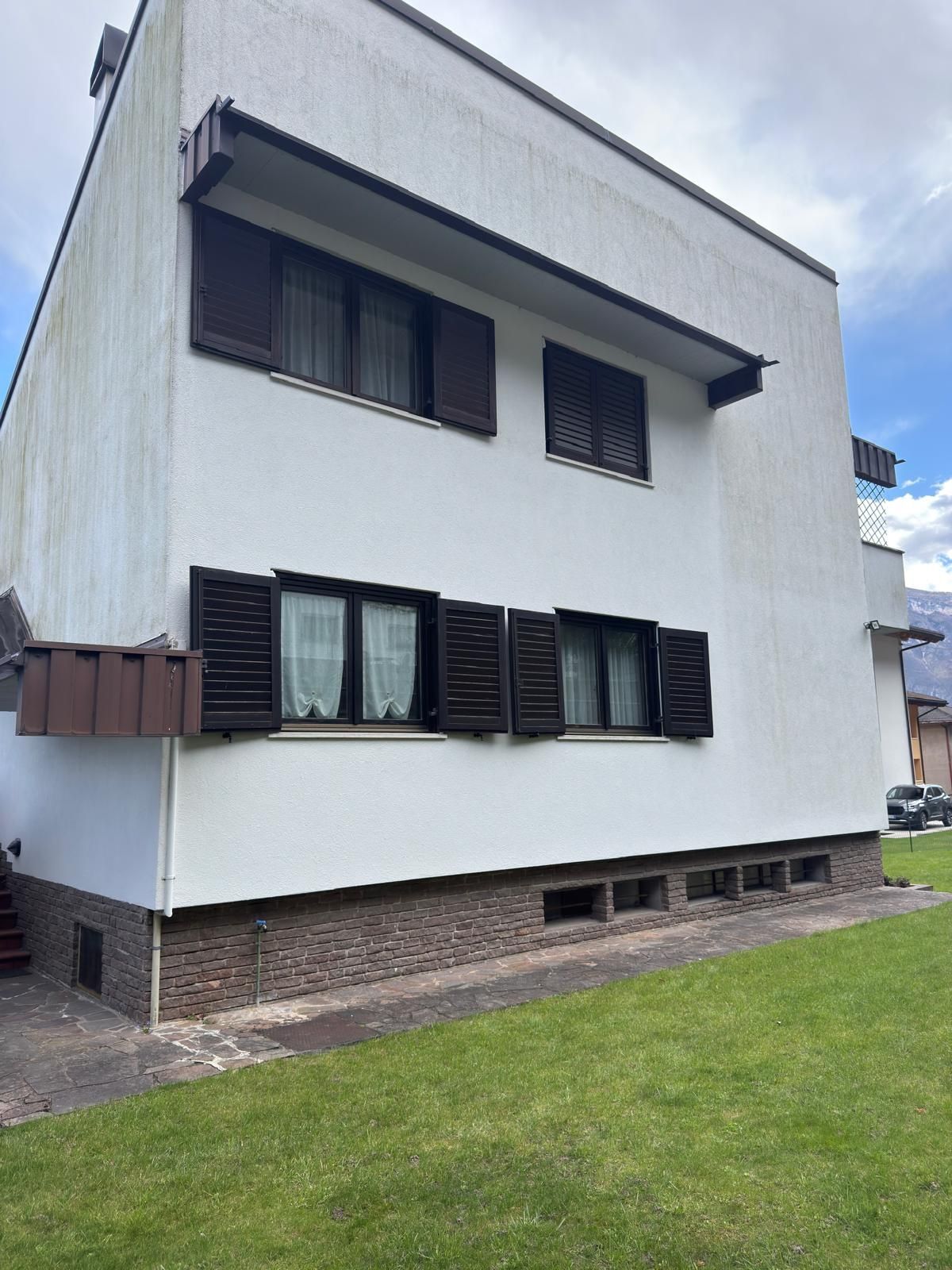 Villa a due piani con facciata chiara, scuri in legno scuro e zoccolatura in mattoni, ripresa dal prato del giardino