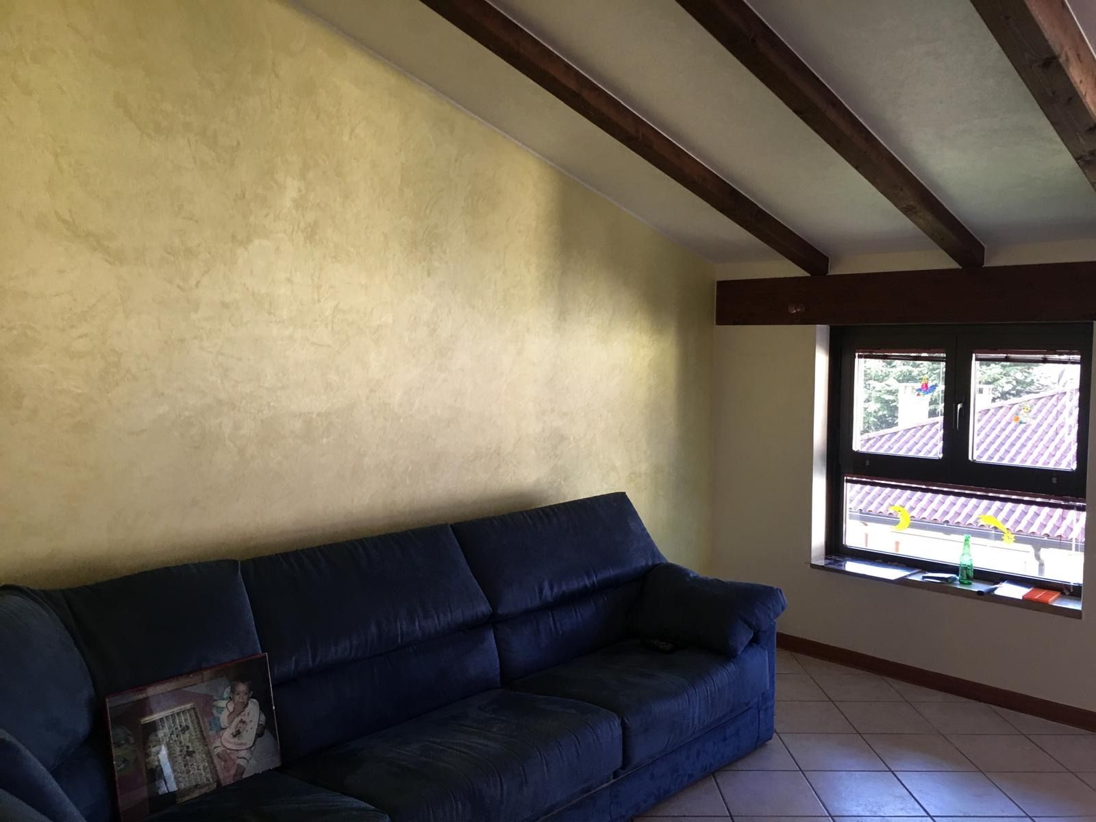 Soggiorno sottotetto con parete decorata a velatura dorata, travi in legno scuro a vista e finestra affacciata sui tetti