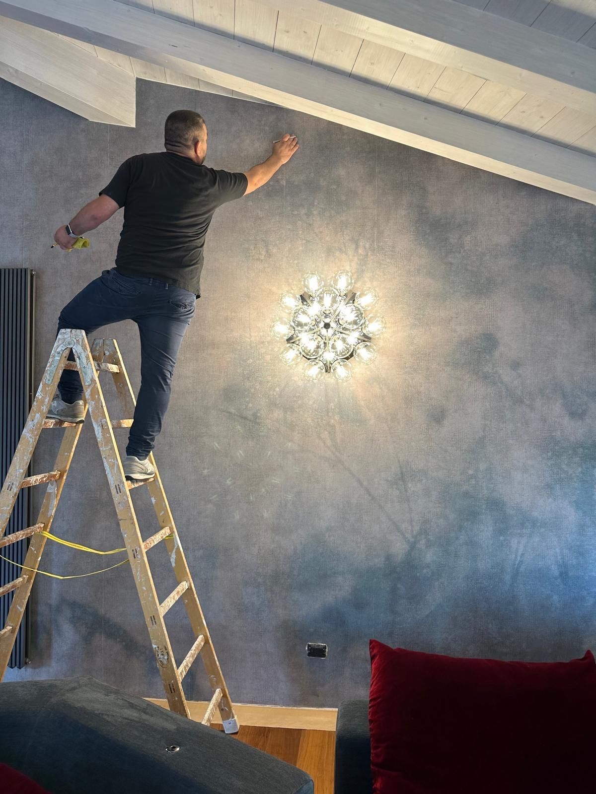 Un decoratore di Enea Pittura, su una scala, rifinisce a mano una parete effetto nuvolato grigio-azzurro in un soggiorno con travi bianche a vista.