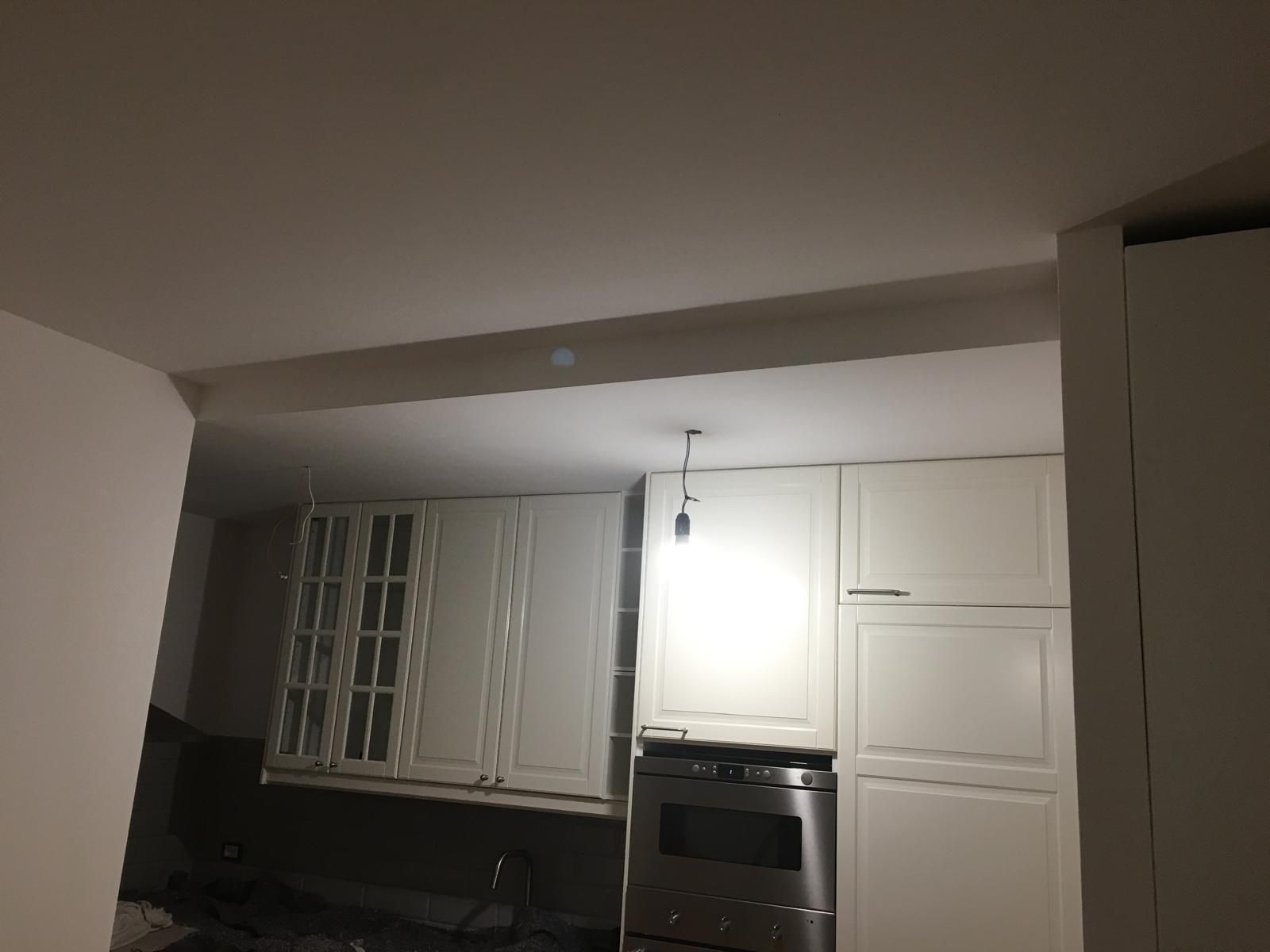 Cucina bianca con pensili, forno a incasso e soffitto ribassato appena tinteggiati, ripresa con la lampadina provvisoria accesa
