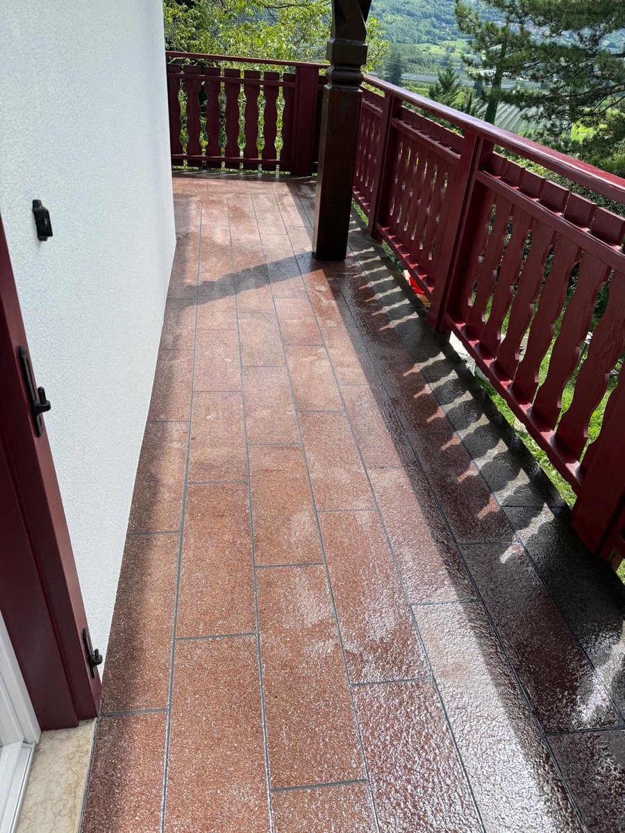 Balcone con parete esterna bianca, ringhiera in legno bordeaux e vista su prati e frutteti