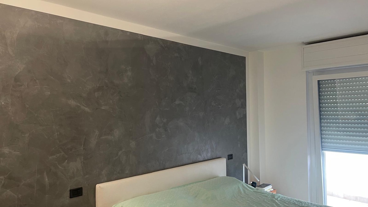 Camera da letto con parete decorativa effetto spatolato grigio dietro la testiera e pareti bianche
