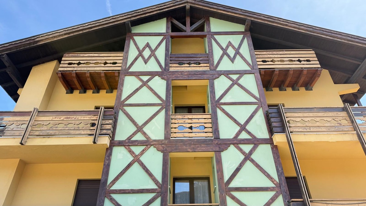 Facciata di edificio alpino tinteggiata giallo e verde menta, con travature scure a X e balconi in legno