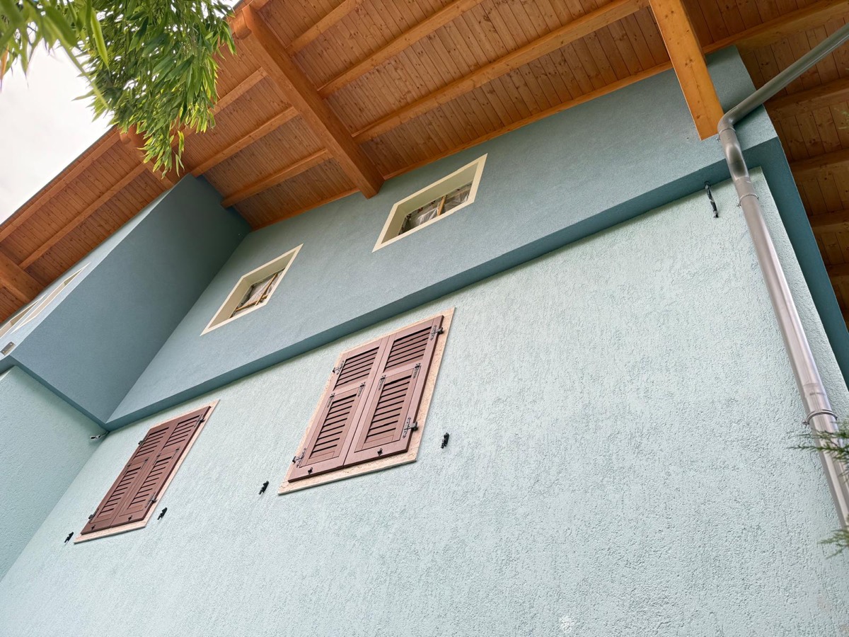 Parete esterna tinteggiata verde salvia con scuri color mattone e travi del tetto in legno chiaro