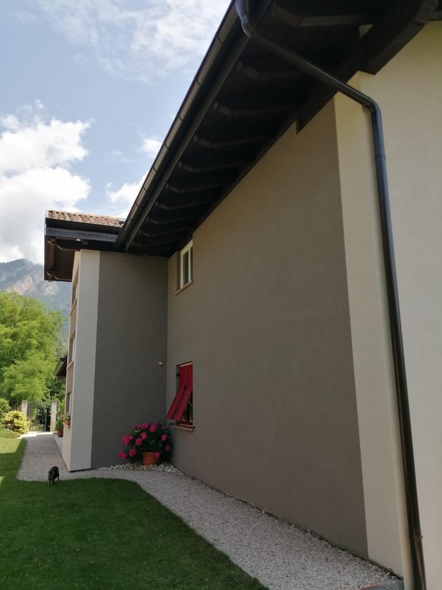 Fianco di casa con parete tinteggiata tortora, sporto in legno scuro, prato e montagna sul fondo