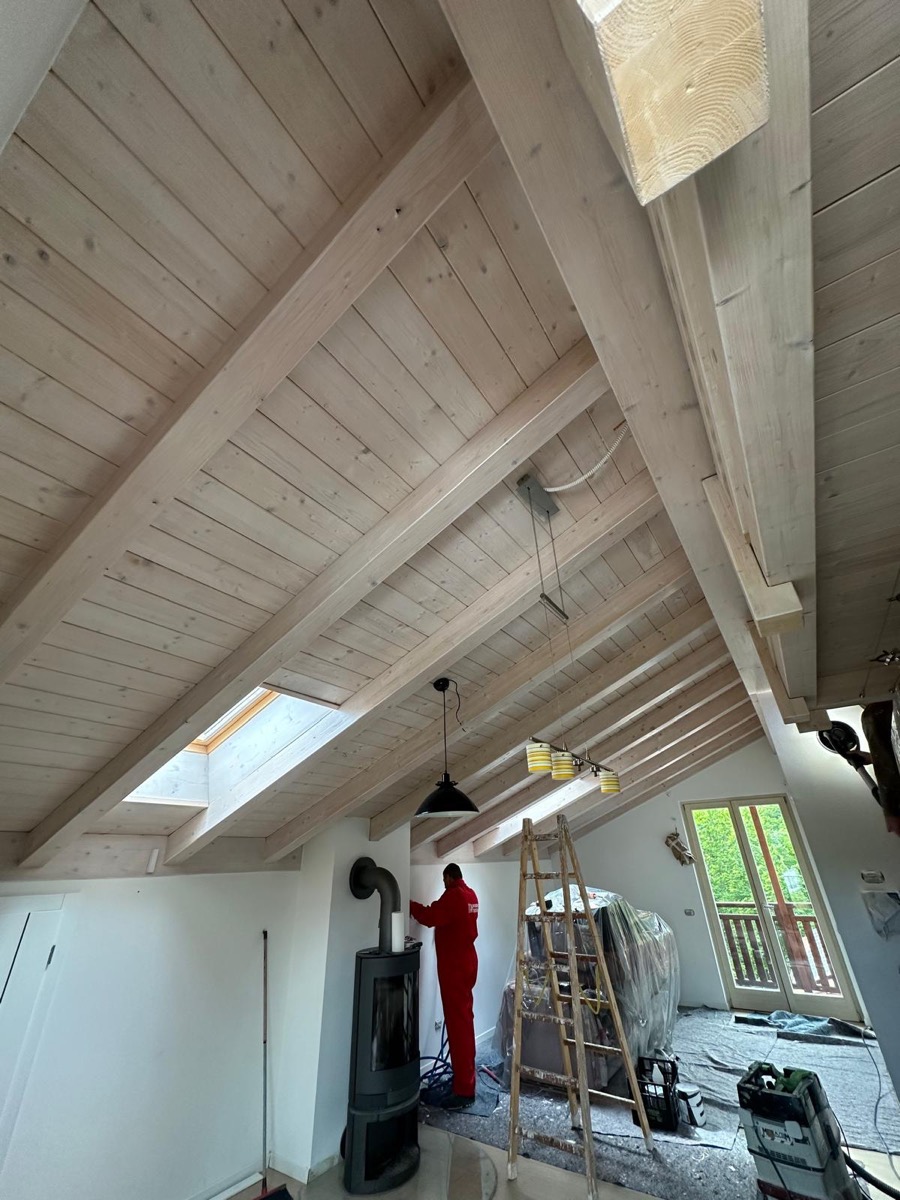 Mansarda con soffitto in legno chiaro a vista: operaio in tuta rossa al lavoro sulla parete