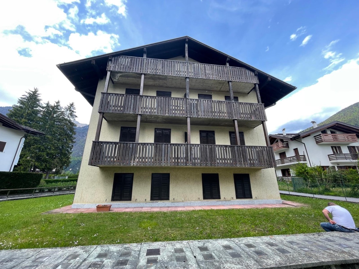 Facciata di una palazzina di montagna con intonaco giallo, ballatoi in legno scuro, prato davanti e montagne sul fondo