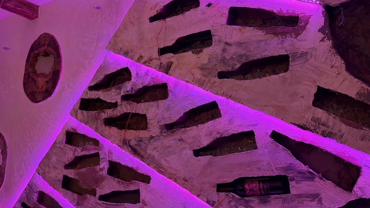 Parete di cantina con nicchie sagomate per bottiglie, finitura materica chiara e illuminazione LED viola