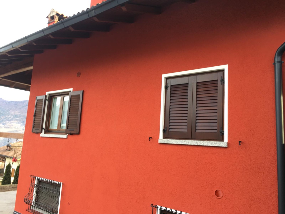 Parete esterna di una casa tinteggiata rosso mattone, con scuri in legno marrone, contorni bianchi alle finestre e davanzali in pietra