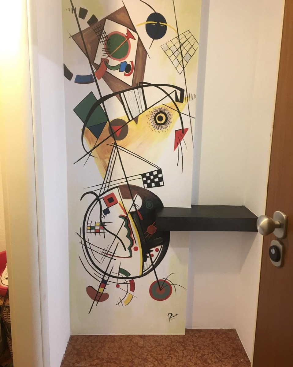 Parete di ingresso con decorazione murale astratta dipinta a mano a colori vivaci, accanto a una mensola nera