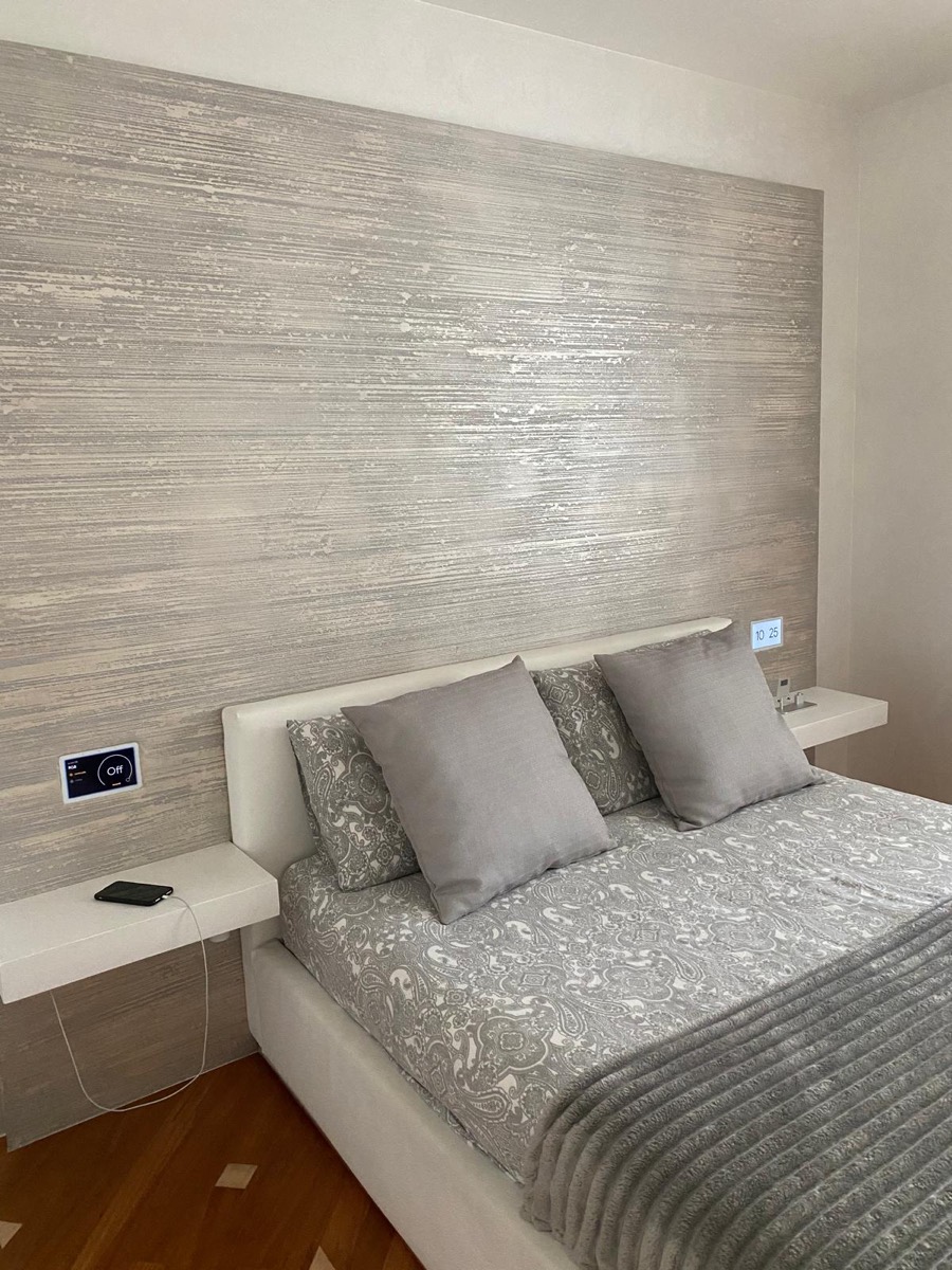 Camera da letto con parete decorata a righe orizzontali effetto perlato dietro un letto imbottito bianco con cuscini grigi