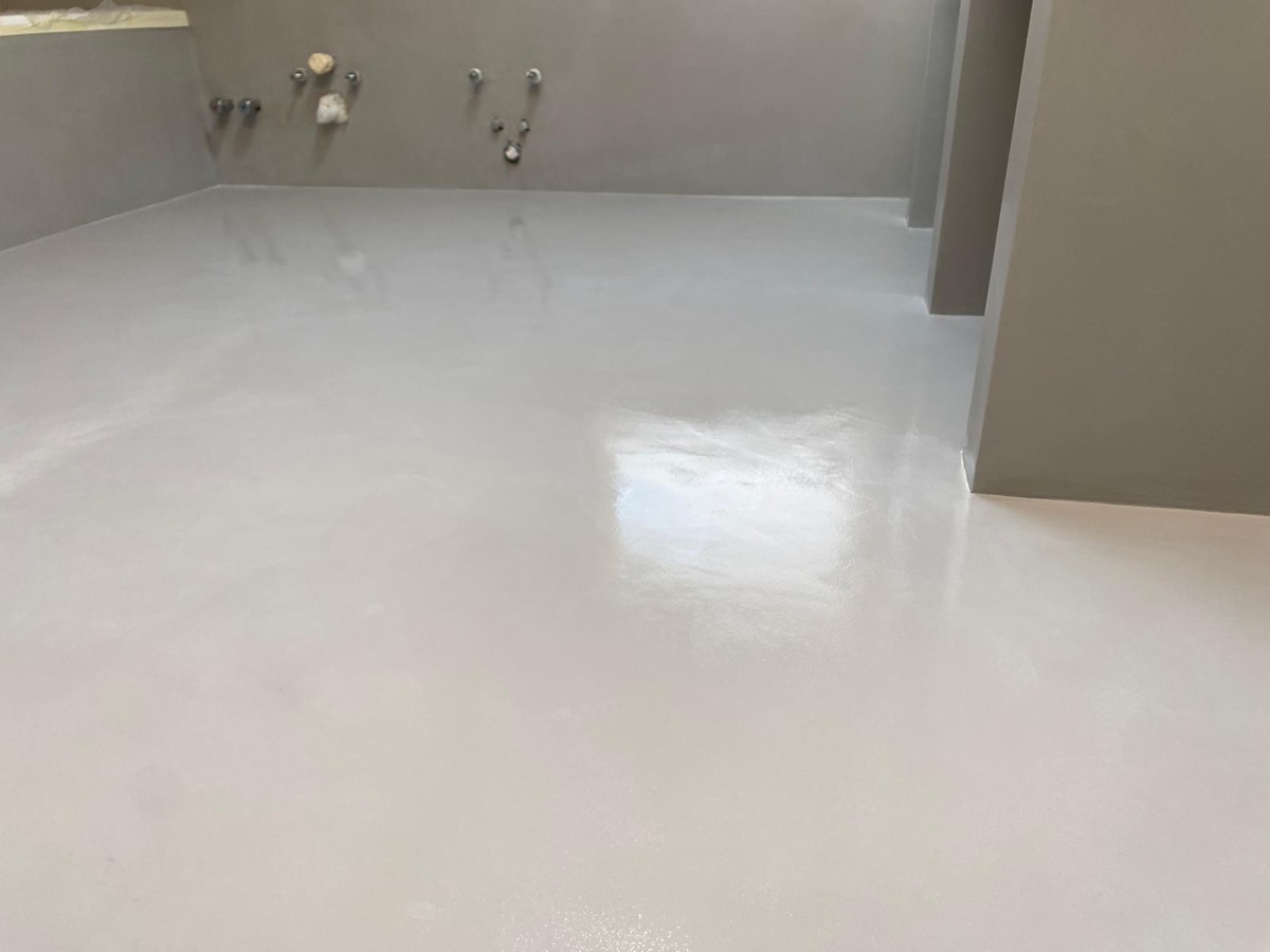 Pavimento in finitura continua chiara appena posato in un bagno con pareti color tortora e attacchi idraulici a vista