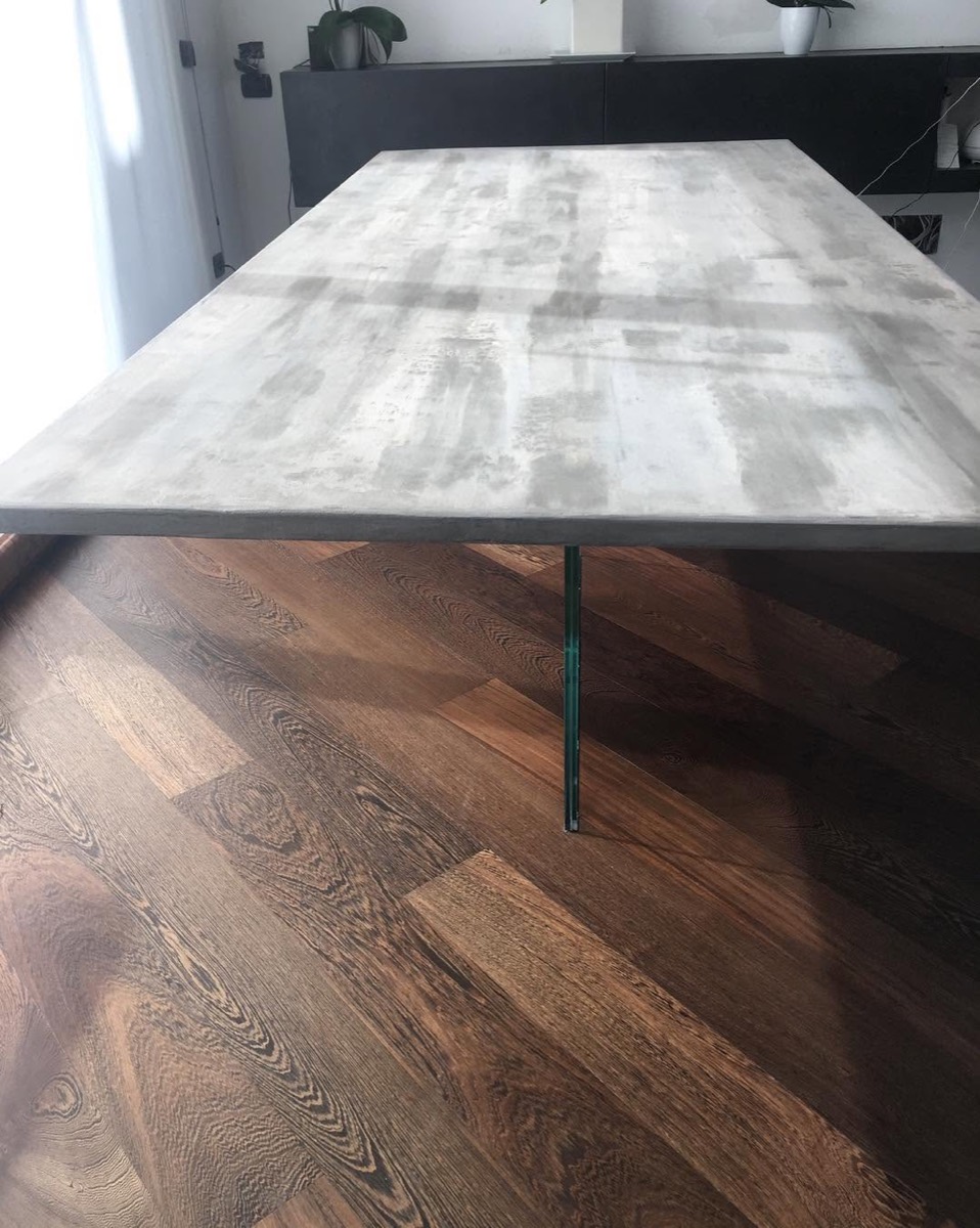 Piano di un tavolo da pranzo rivestito in microcemento grigio effetto cemento, con gamba in vetro, su pavimento in parquet di rovere scuro