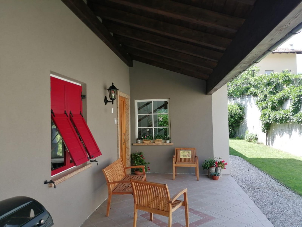 Portico di casa con pareti tinteggiate tortora, persiane rosse, mobili in legno e giardino