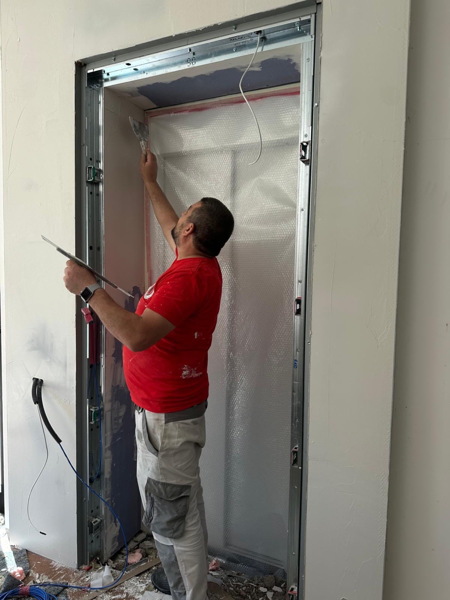 Operaio rasa con la spatola la spalletta di un vano porta con telaio metallico e telo di protezione