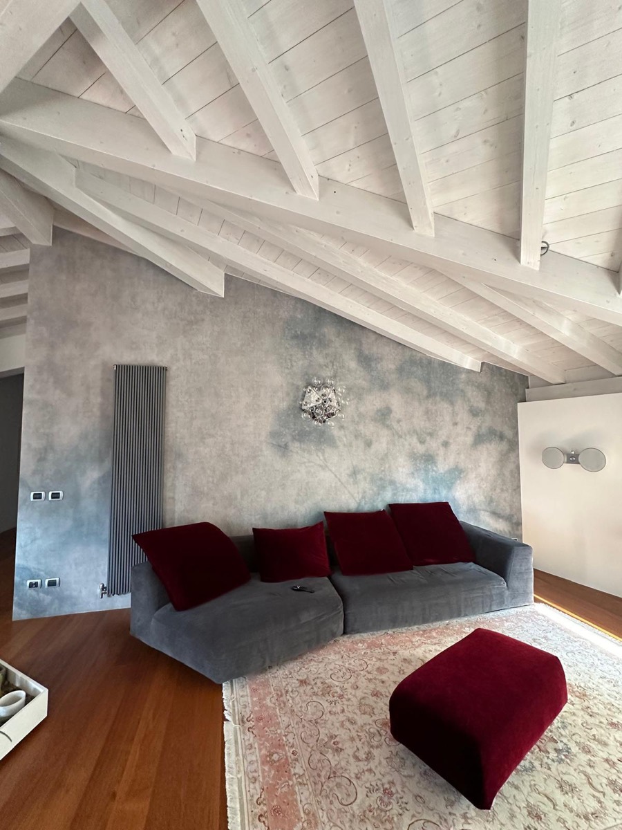 Soggiorno sottotetto con travi in legno bianche e parete decorativa effetto nuvolato grigio-azzurro dietro il divano