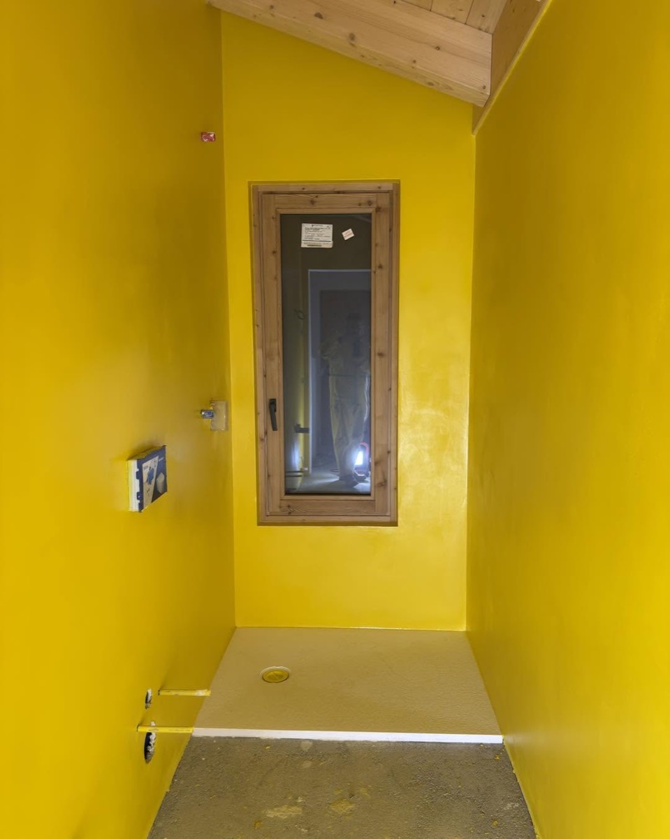 Vano doccia in cantiere con guaina liquida impermeabilizzante gialla stesa su pareti e piatto doccia, finestra in legno e travi a vista