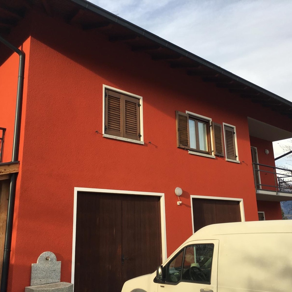 Facciata di un'abitazione intonacata in rosso-arancio con persiane e portoni in legno