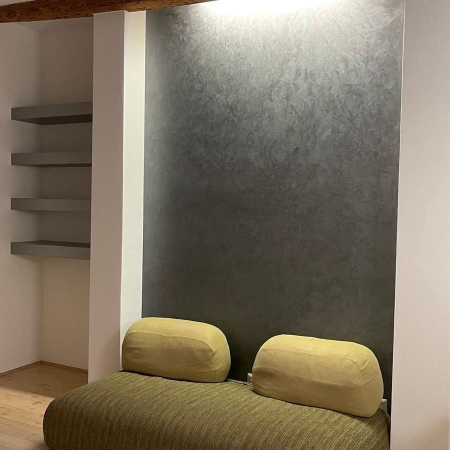Soggiorno con parete decorativa effetto spatolato grigio antracite, mensole a incasso e divano verde