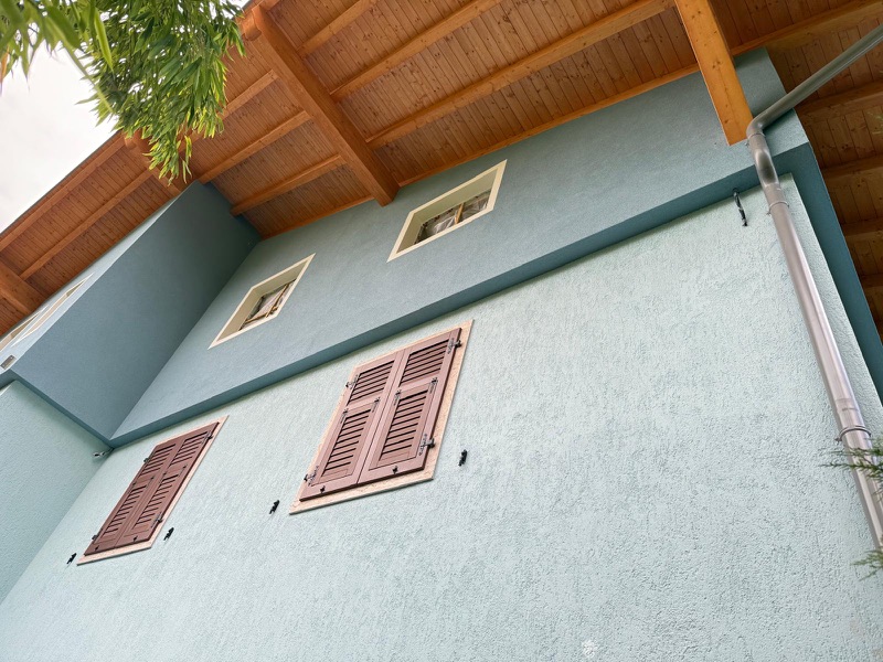 Parete esterna tinteggiata verde salvia con scuri color mattone e travi del tetto in legno chiaro