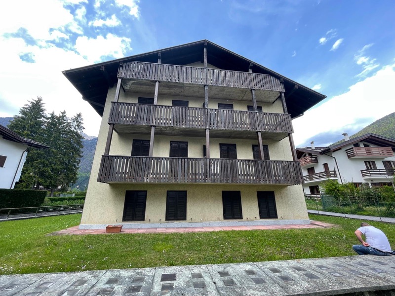 Facciata di una palazzina di montagna con intonaco giallo, ballatoi in legno scuro, prato davanti e montagne sul fondo