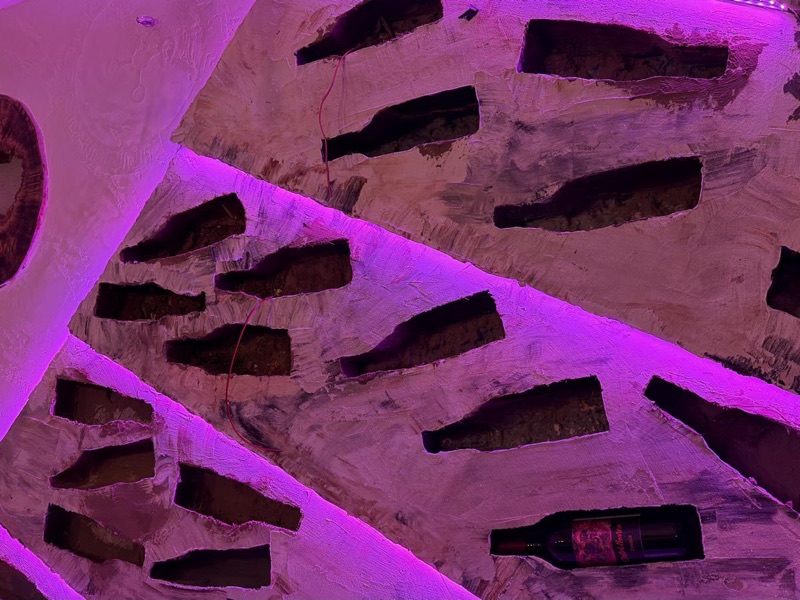 Parete di cantina con nicchie sagomate per bottiglie, finitura materica chiara e illuminazione LED viola